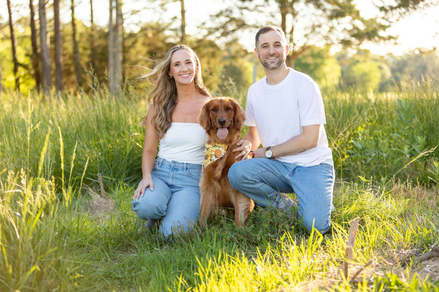 A couple kneeling in a sunlit meadow with their brown dog during golden hour.