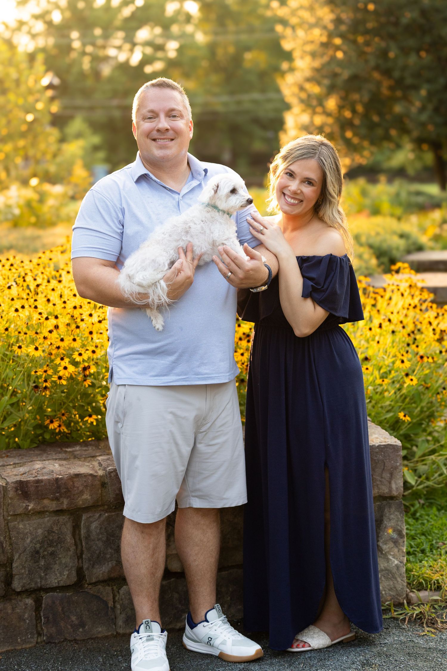 Smiling couple holding a small white dog outdoors with yellow flowers and green trees in the background.