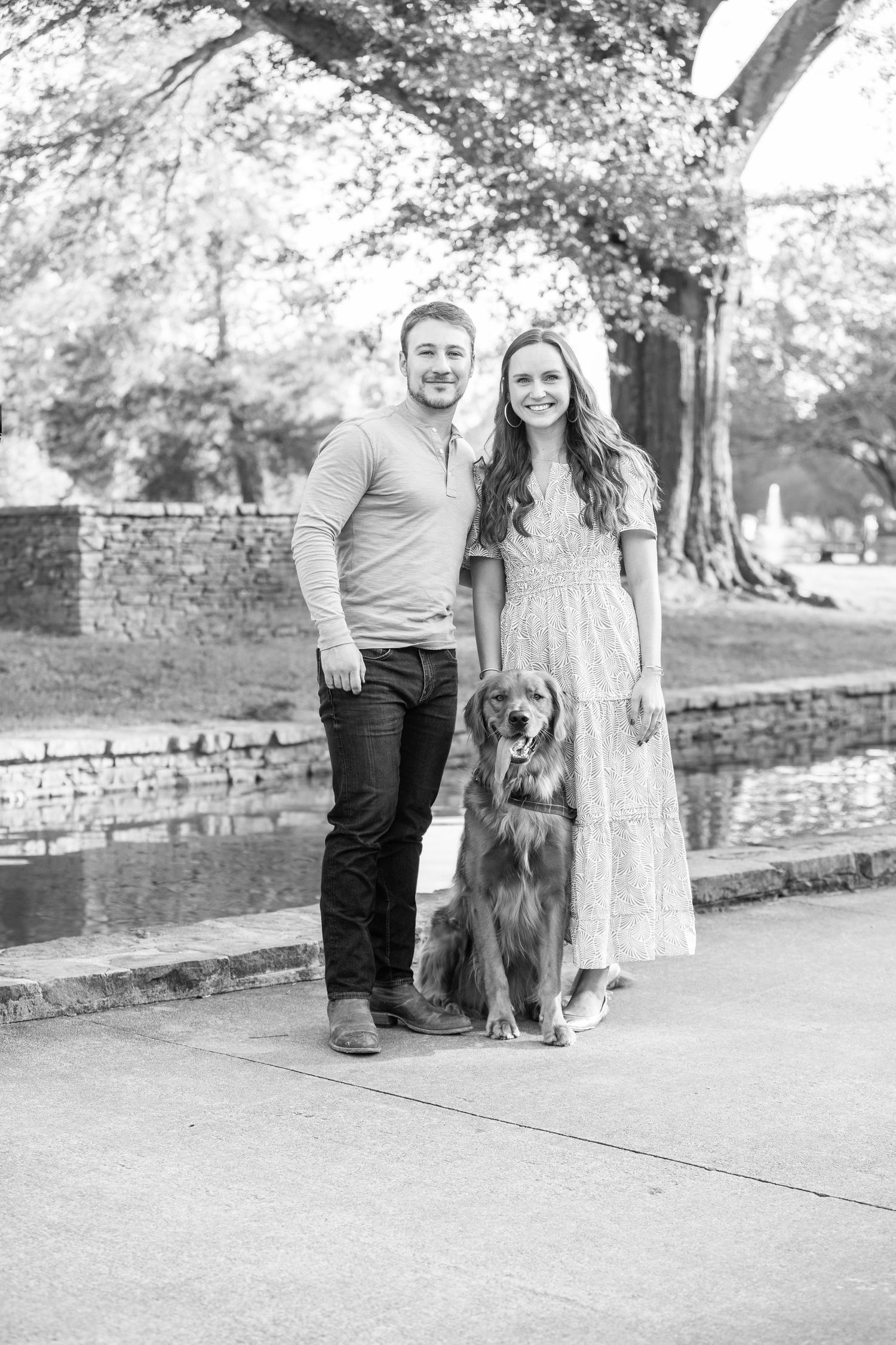 A couple stands with their golden retriever dog on a stone-lined path in a park, captured in black and white.