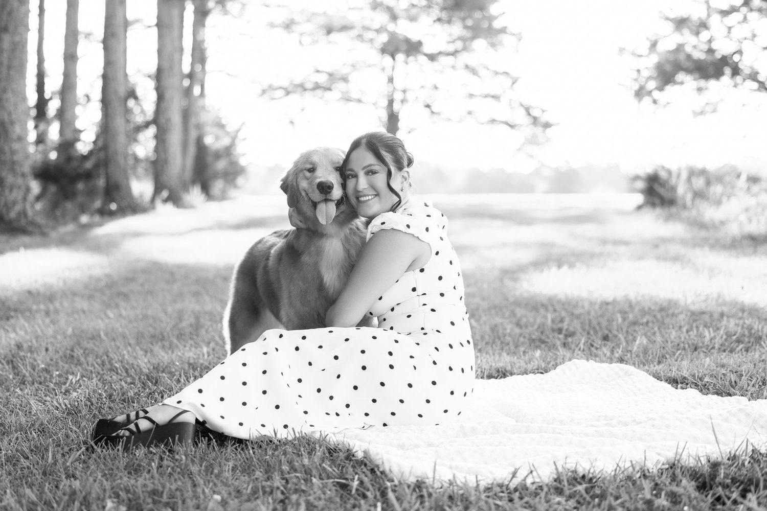 Woman in polka dot dress hugging a golden retriever dog while sitting on grass in a black and white photo.