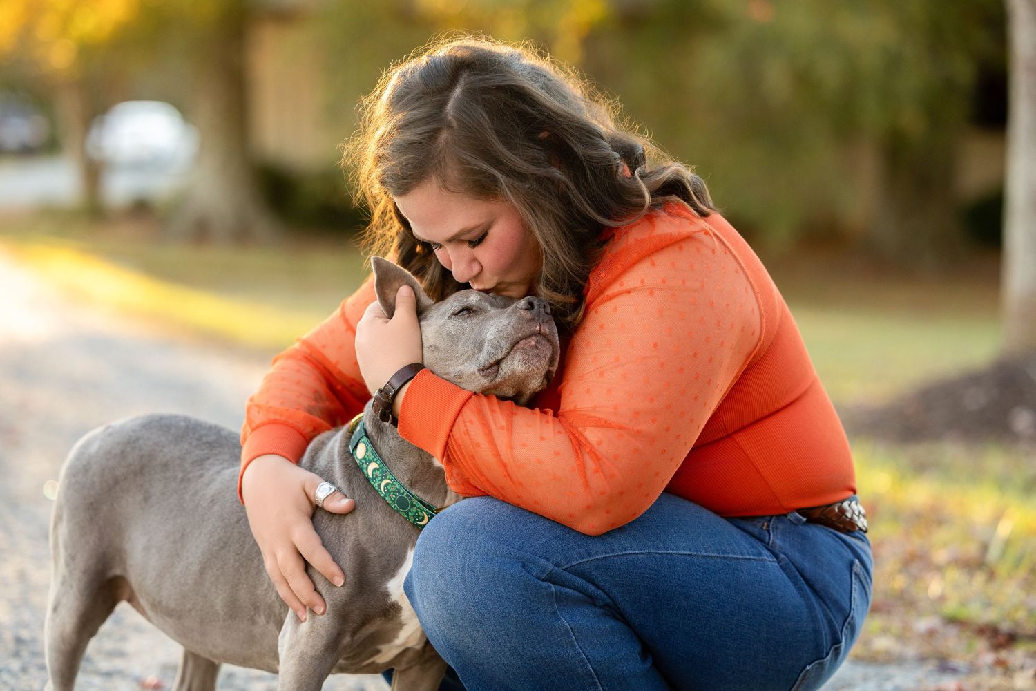 A person in an orange sweater sits outdoors with a gray Weimaraner dog during autumn.