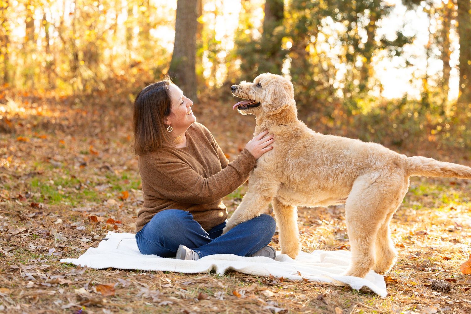 Woman sitting on blanket in autumn forest petting a golden doodle dog outdoors.