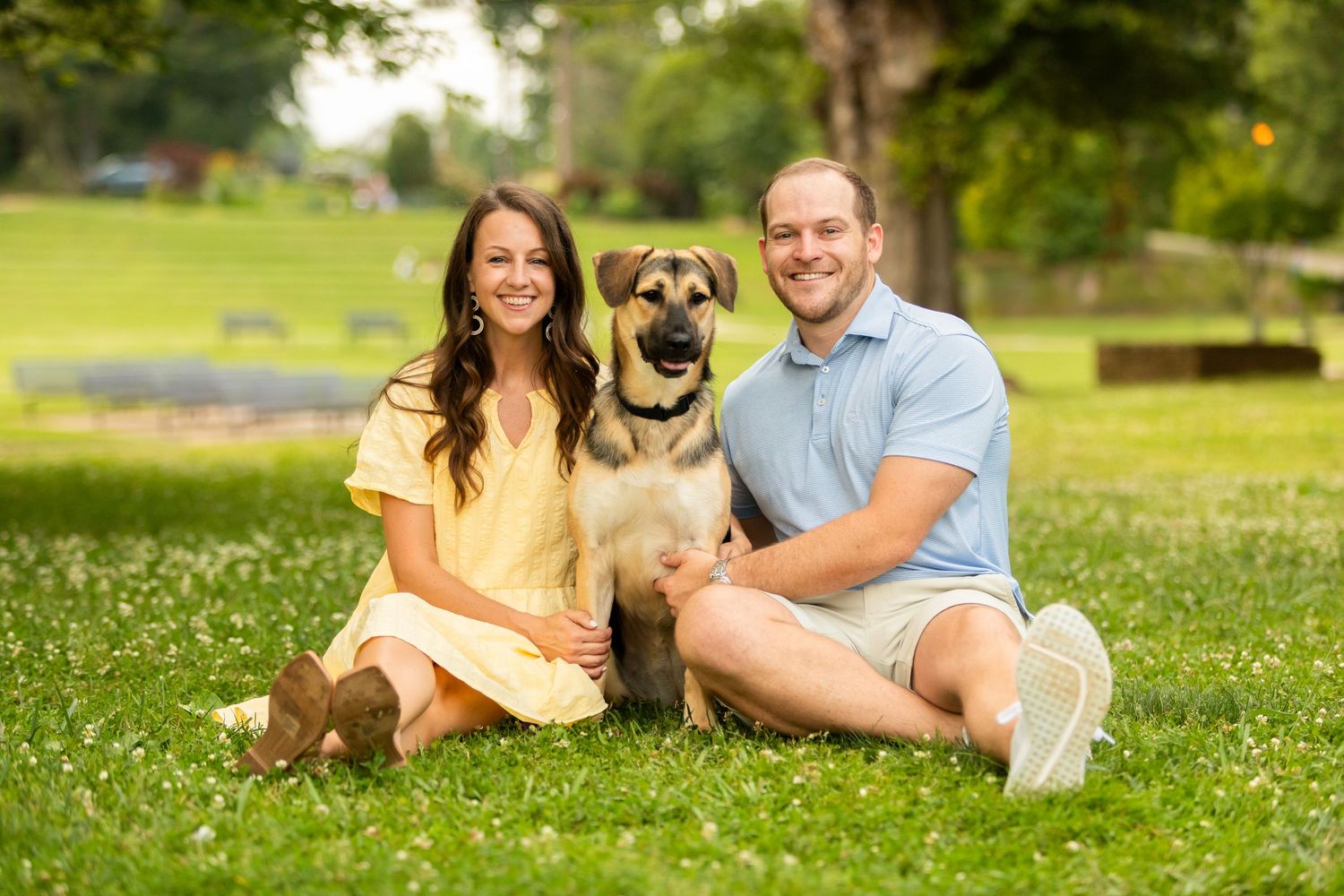 A couple relaxes with their German Shepherd dog on a sunny grassy lawn with a water fountain in the background.