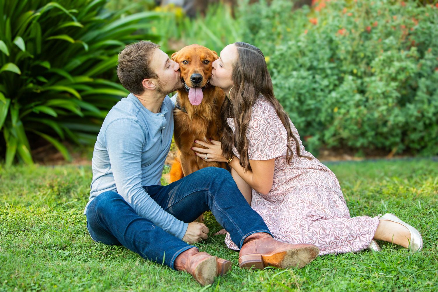 A loving couple kisses their happy golden retriever dog while sitting together on green grass in a garden.