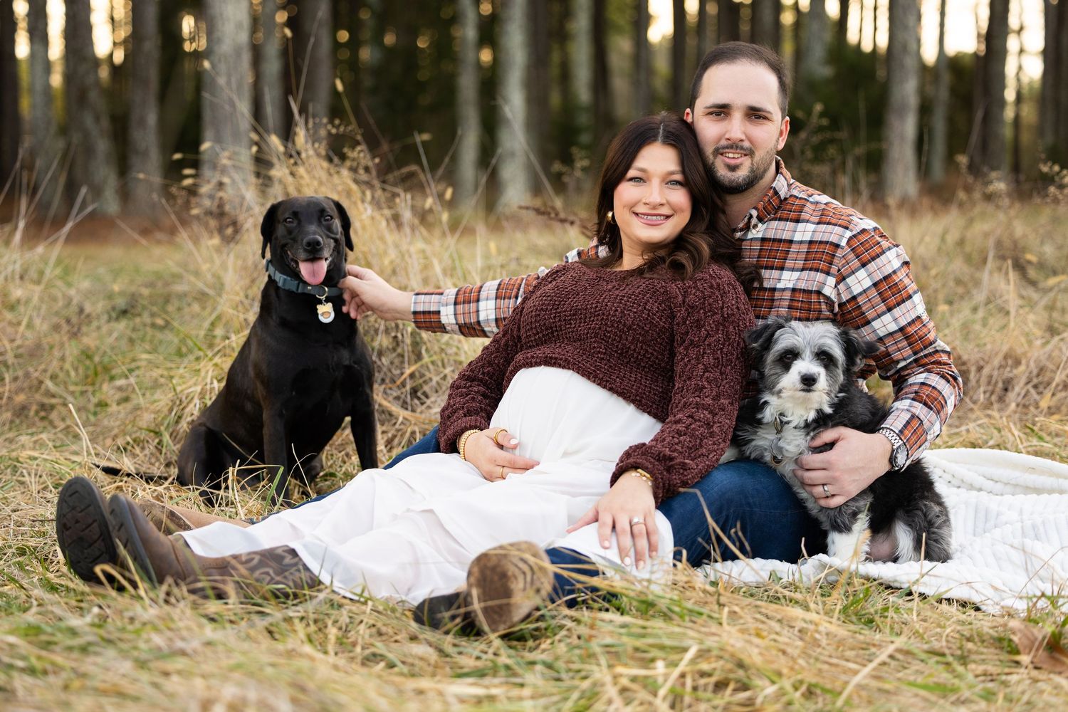 Family and their black dogs pose together on a white blanket in a rustic outdoor setting during autumn.