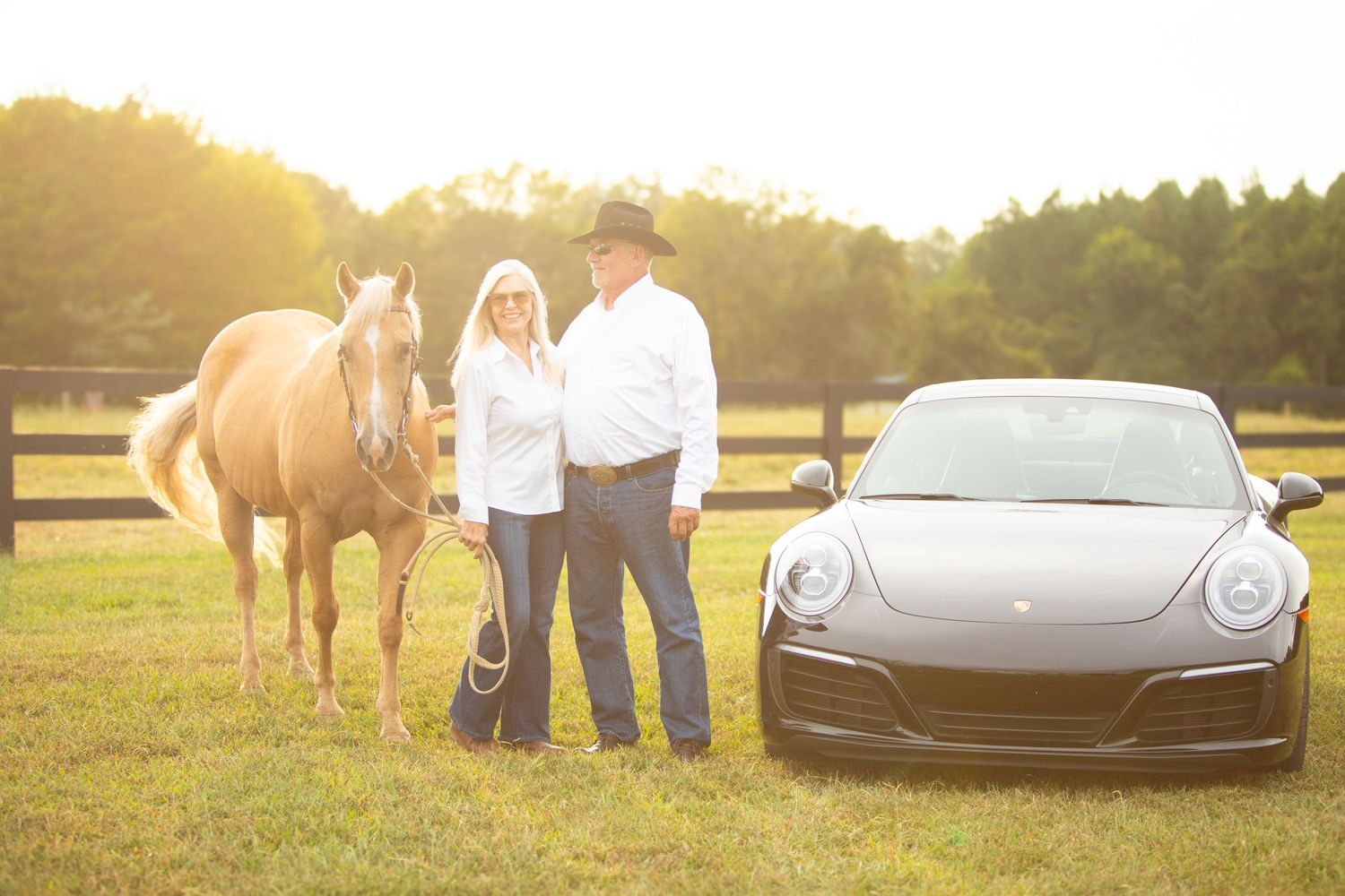 Couple and their palomino horse pose together at sunset with a black Porsche sports car in a grassy field.