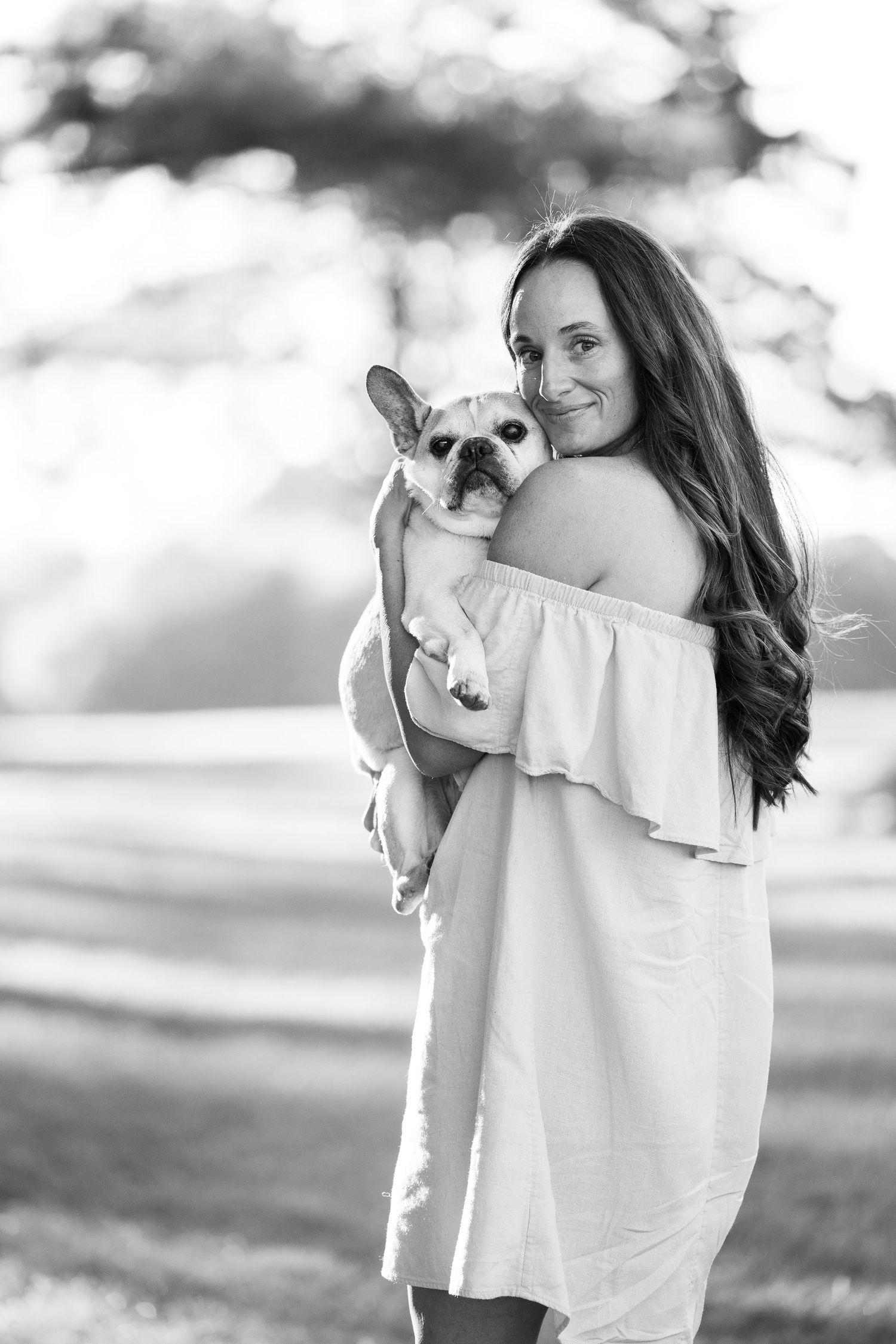 Black and white portrait of woman in off-shoulder dress holding small French Bulldog puppy outdoors.