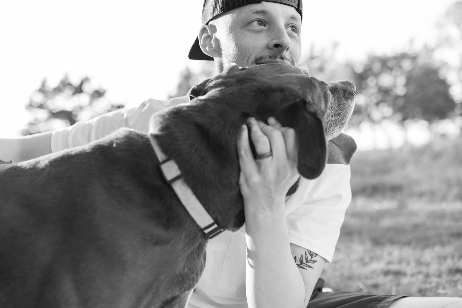 Black and white photo of a man in a cap hugging a dog outdoors in a park setting.
