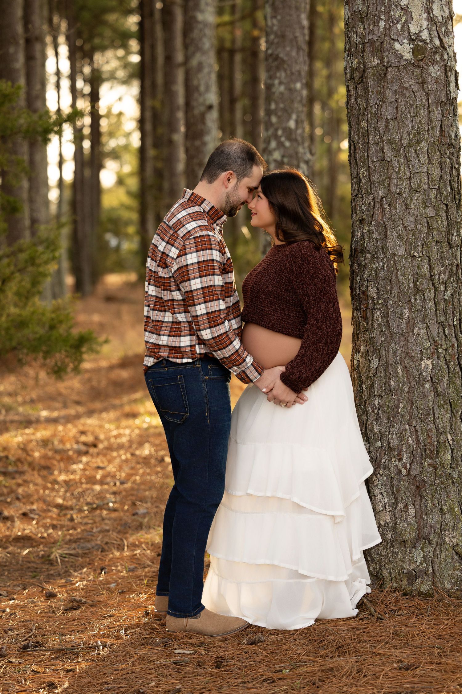 A couple shares an intimate moment among tall pine trees in a rustic forest engagement photo session at sunset.