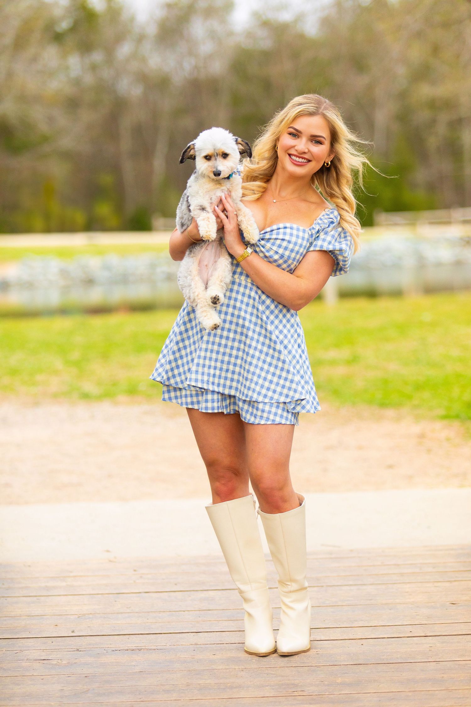 Person wearing blue gingham romper and white boots holds small white puppy while standing on wooden deck outdoors.