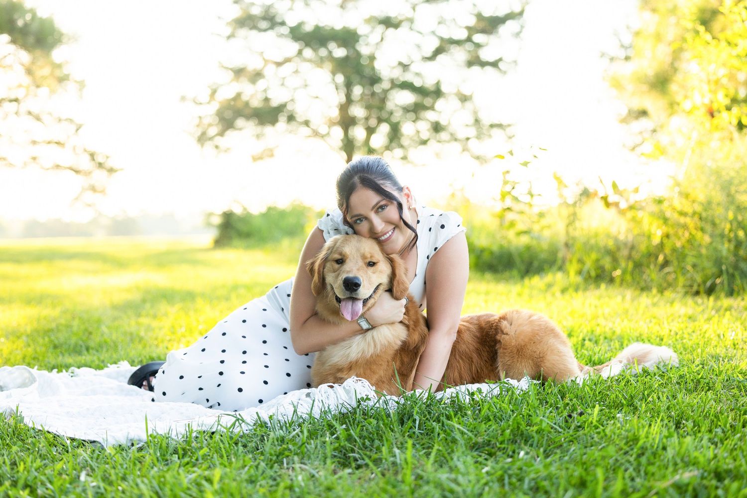 Woman in polka dot dress hugging golden retriever on blanket in sunny green field.