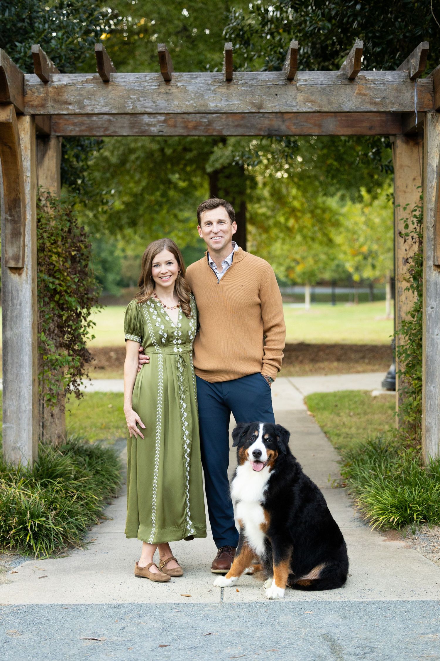 A smiling couple in a green dress and tan sweater stand arm in arm with a Bernese Mountain Dog beneath a wooden pergola in a park.