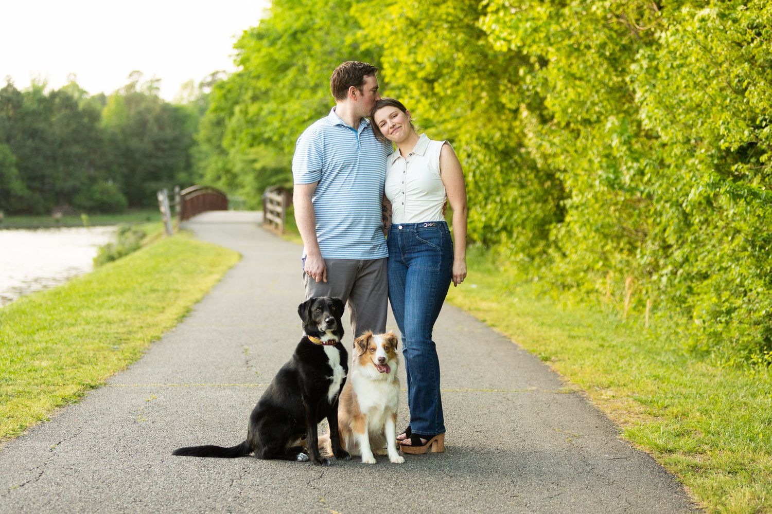 A couple walks their two dogs, a black lab and a gold retriever mix, on a sunny path near water and green trees.