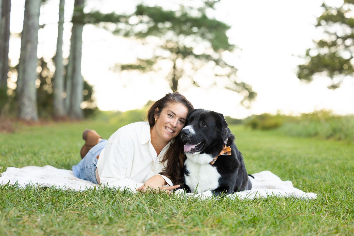A person in white lies on grass with a black and white Border Collie dog during an outdoor photo session.