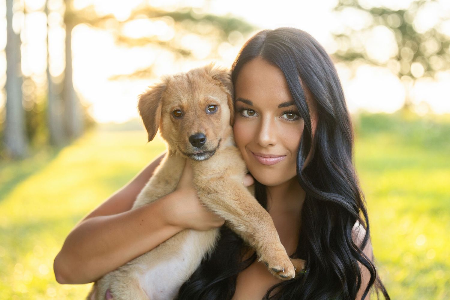 Young woman with long dark hair cuddling a cute brown puppy in a sunlit outdoor setting.