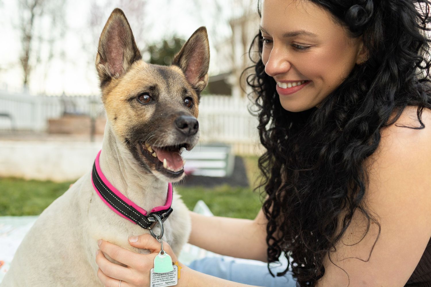 Woman with curly hair smiling at her happy dog wearing a pink collar outdoors.