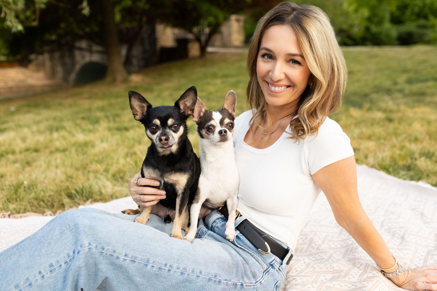 Woman smiling outdoors on blanket with two Chihuahua dogs in a sunny park setting.