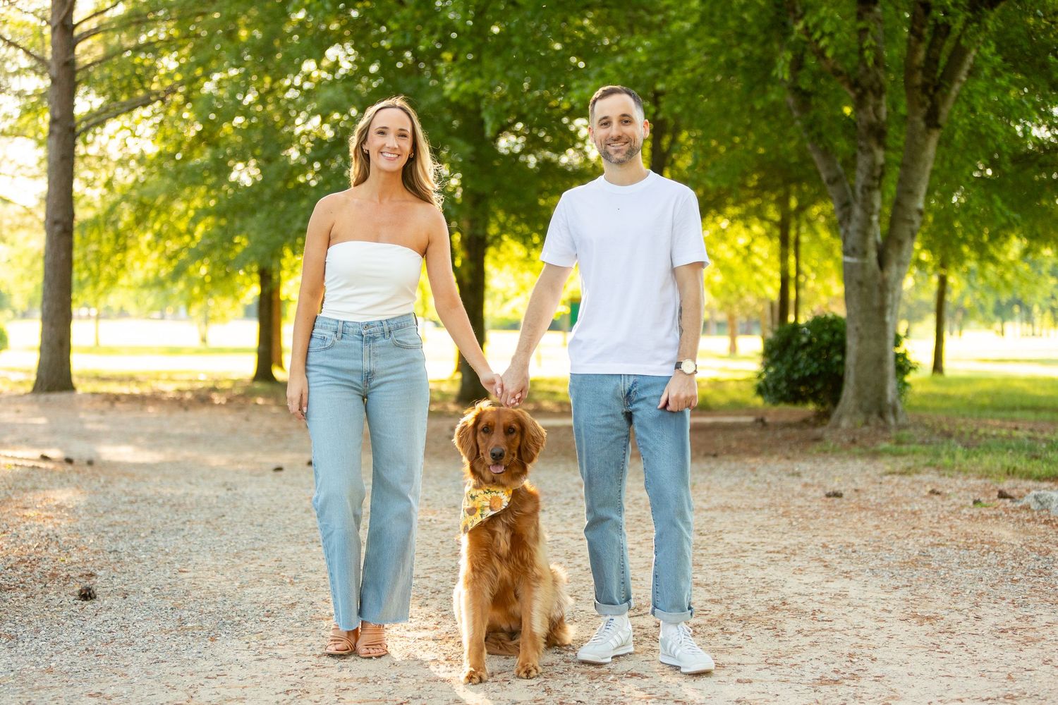 A golden retriever dog walks on a tree-lined path with two people in jeans during a sunny afternoon.