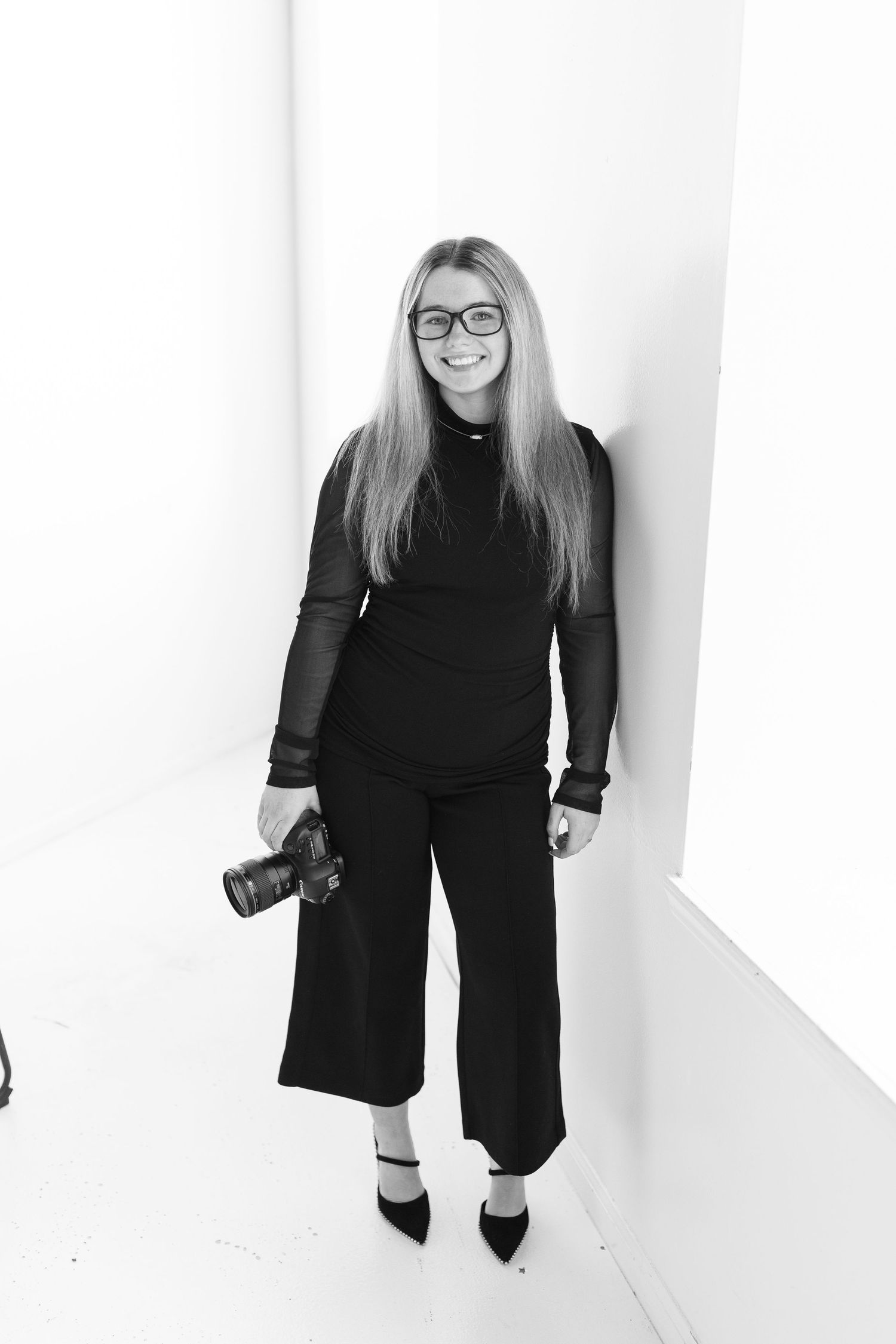 Professional photographer in black outfit holding camera against white background.