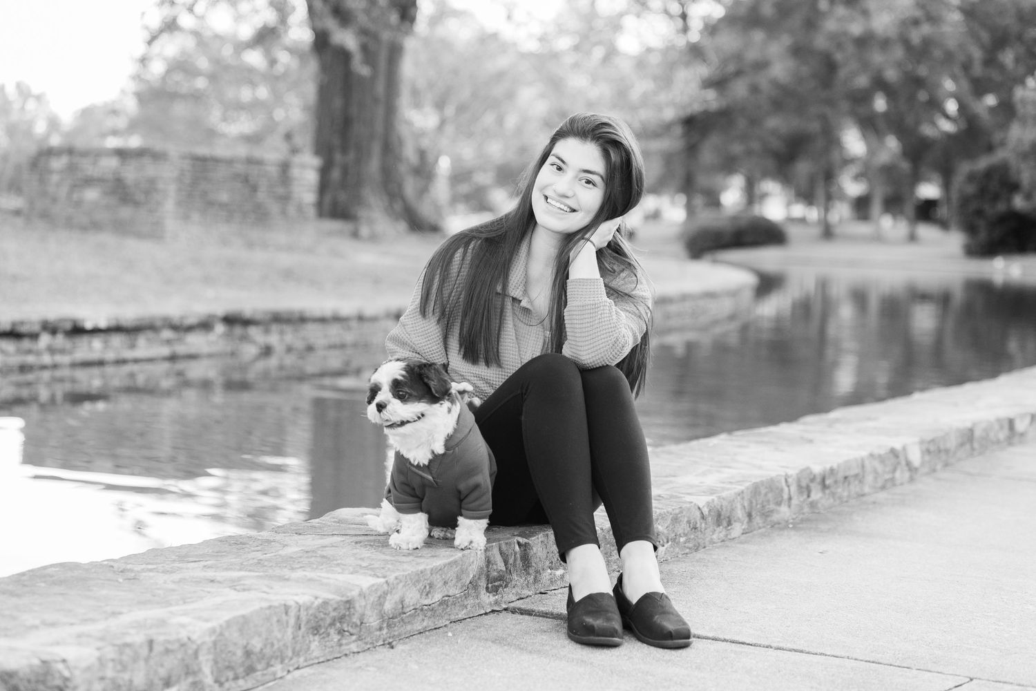 Woman and small dog in sweater sit together on stone wall in black and white portrait.