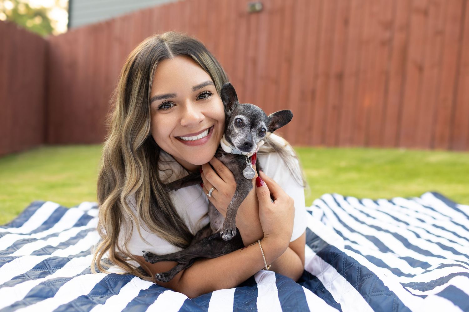 Smiling young woman lying on striped blanket outdoors, hugging a small black and white Chihuahua dog.