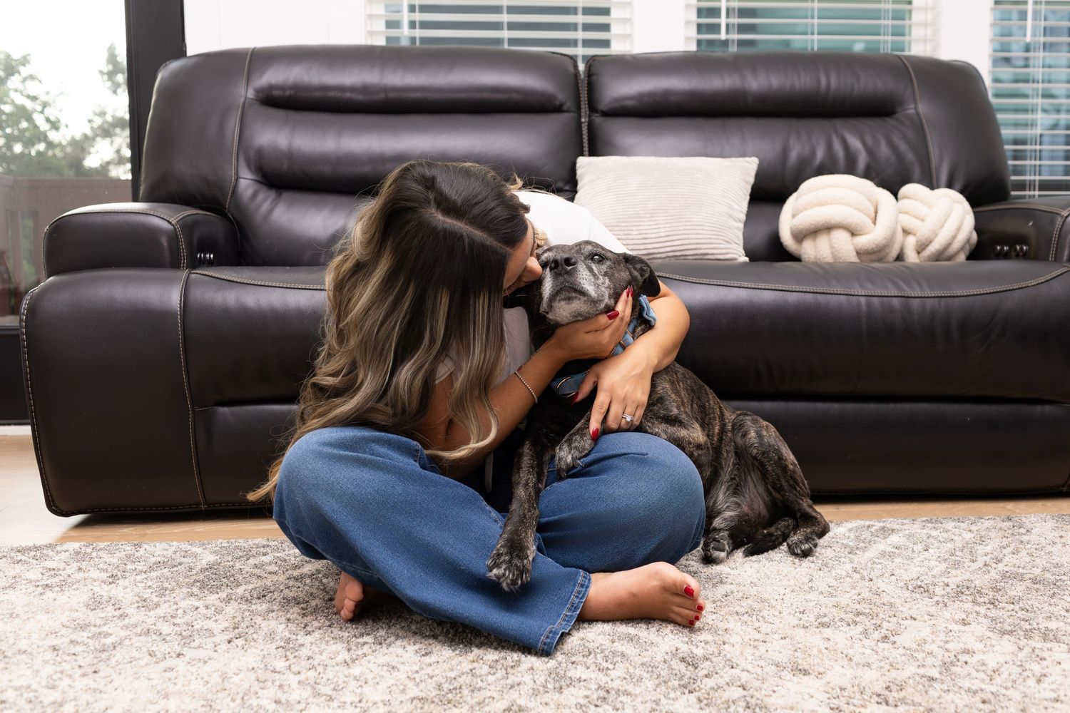 Woman sitting cross-legged on rug kissing her small black dog in front of a black leather sofa.