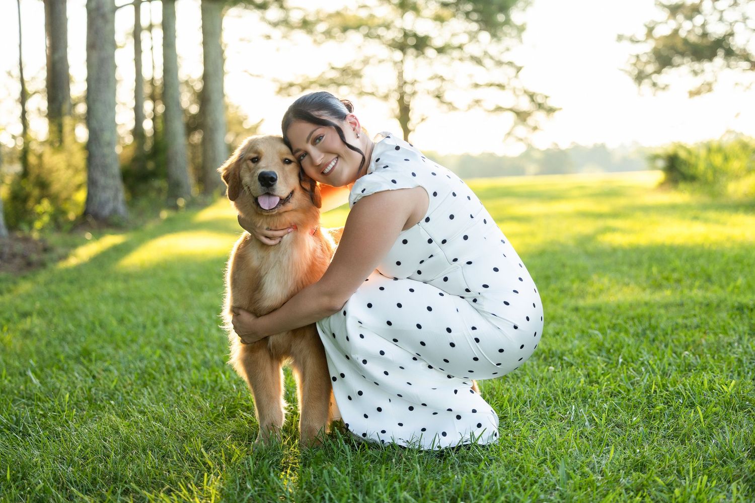 Woman in polka dot dress hugging golden retriever dog on green grass with sunlit trees in background.