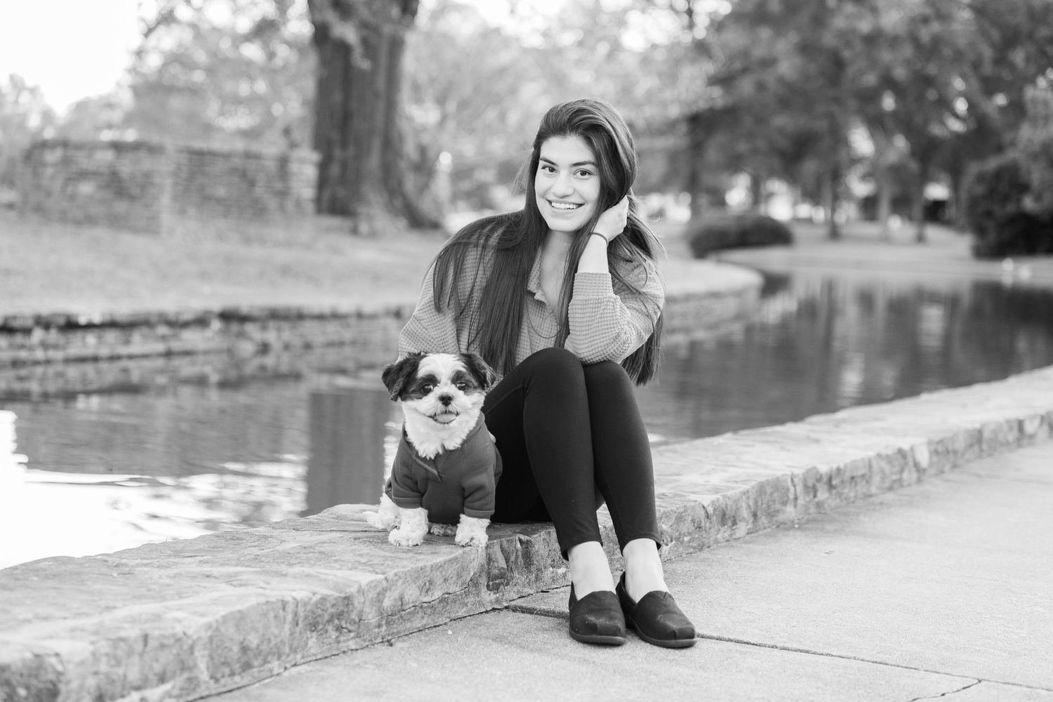 Woman poses casually with small dog on stone wall in black and white photograph.
