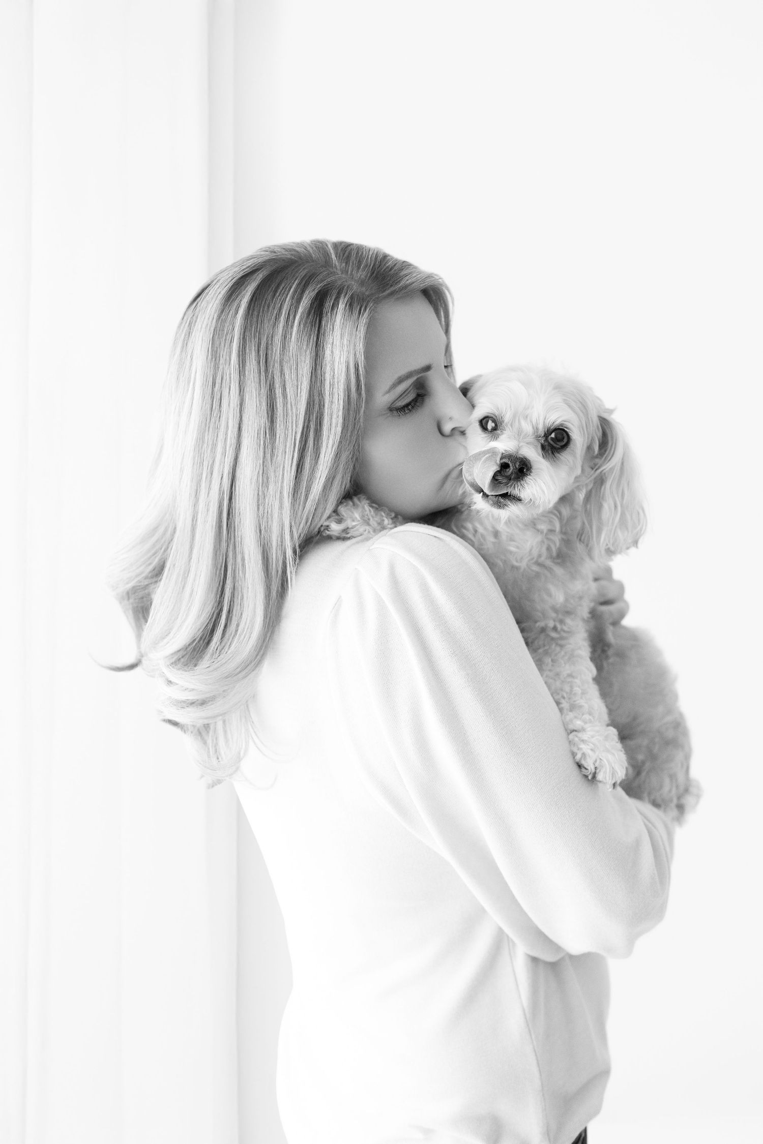 Black and white photo of a blonde woman kissing her small fluffy dog while holding it in her arms.