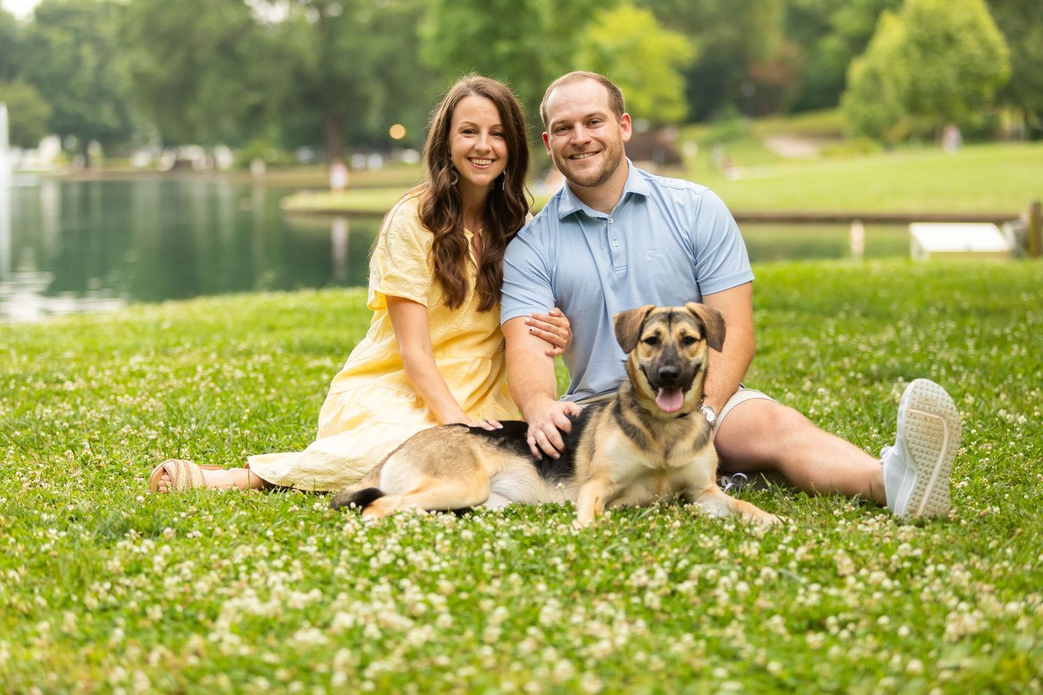 A couple sits on grass by a lake with their brown dog on a sunny summer day.
