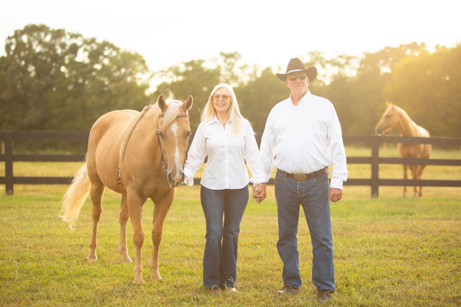 Couple in western attire stand hand-in-hand with their palomino horse in a sunlit pasture.