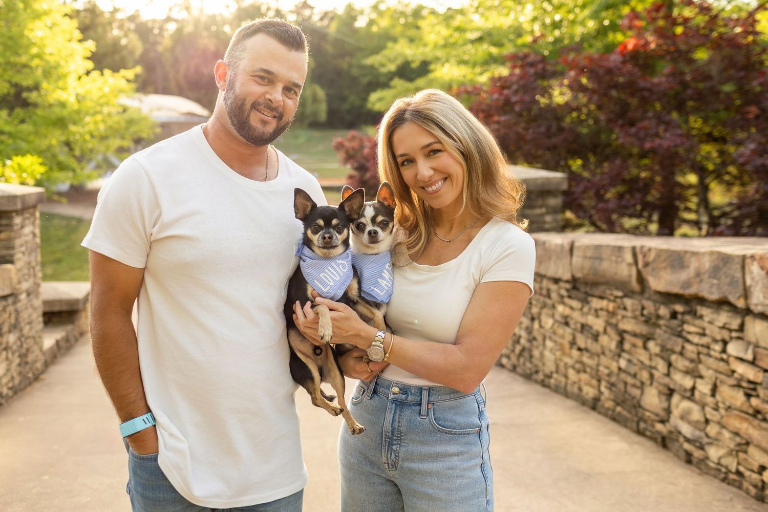Smiling couple in white shirts holding a small Chihuahua in a blue outfit outdoors near a stone wall.