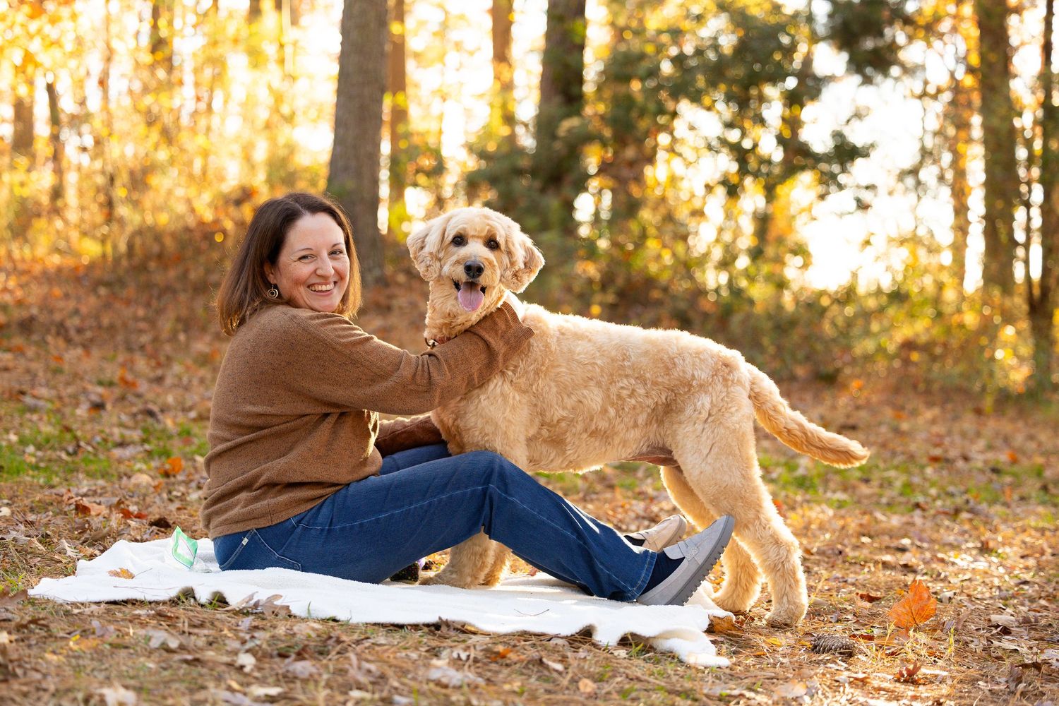 Woman smiling with golden doodle dog on blanket in autumn forest with golden foliage.