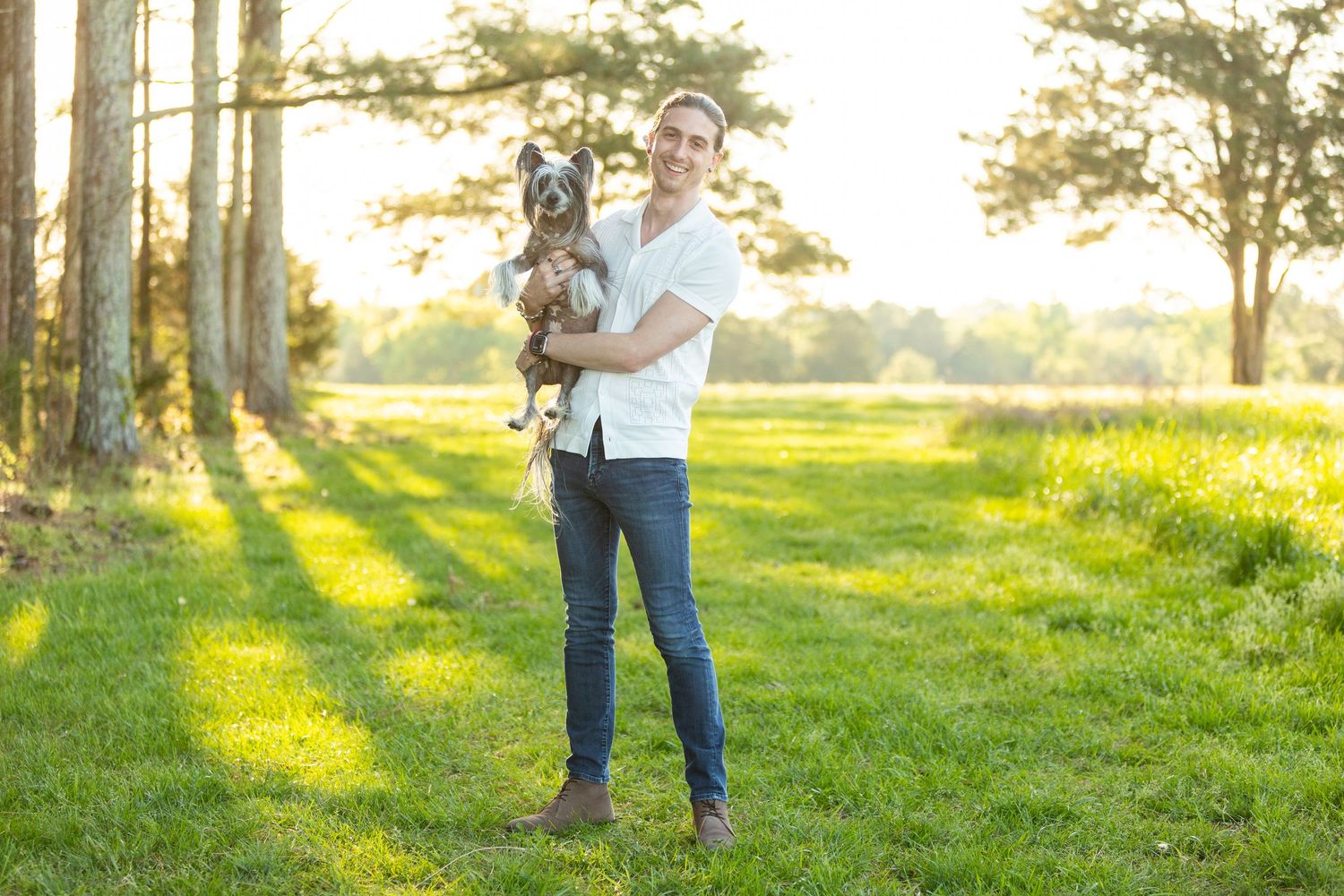 Someone holding a small black dog stands in a sunlit grass field at golden hour with trees in the background.