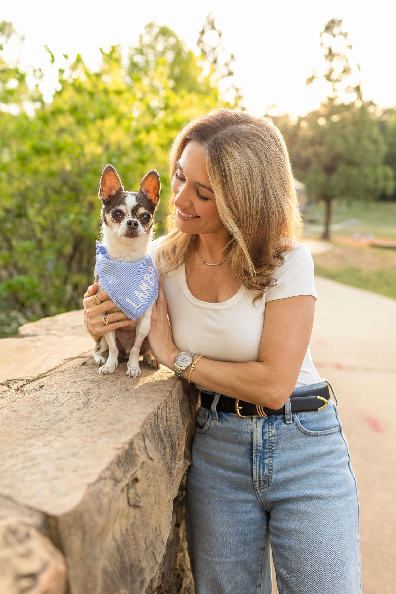 Smiling blonde woman holding a small Chihuahua wearing a blue bandana outdoors in a sunny park setting.