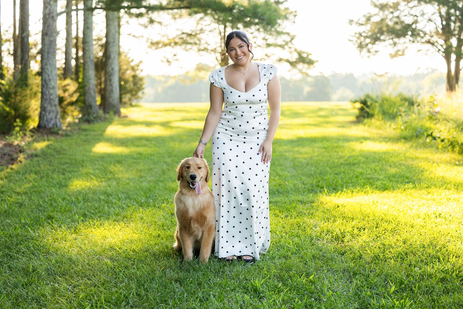 Woman in white polka dot maxi dress poses with golden retriever on lush green lawn surrounded by tall trees.