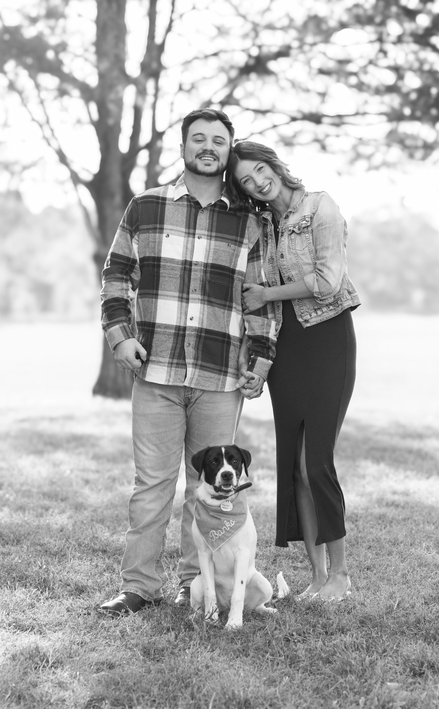Black and white portrait of couple with their dog standing together in a park setting.