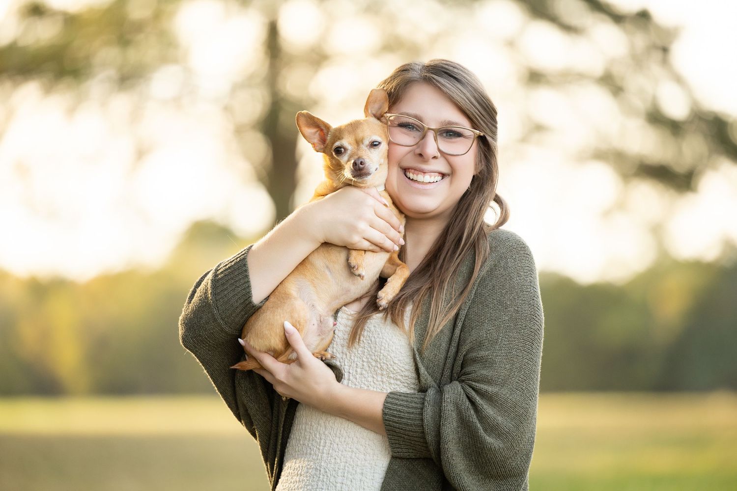A person in a gray sweater holds a small corgi puppy in an outdoor setting during golden hour.