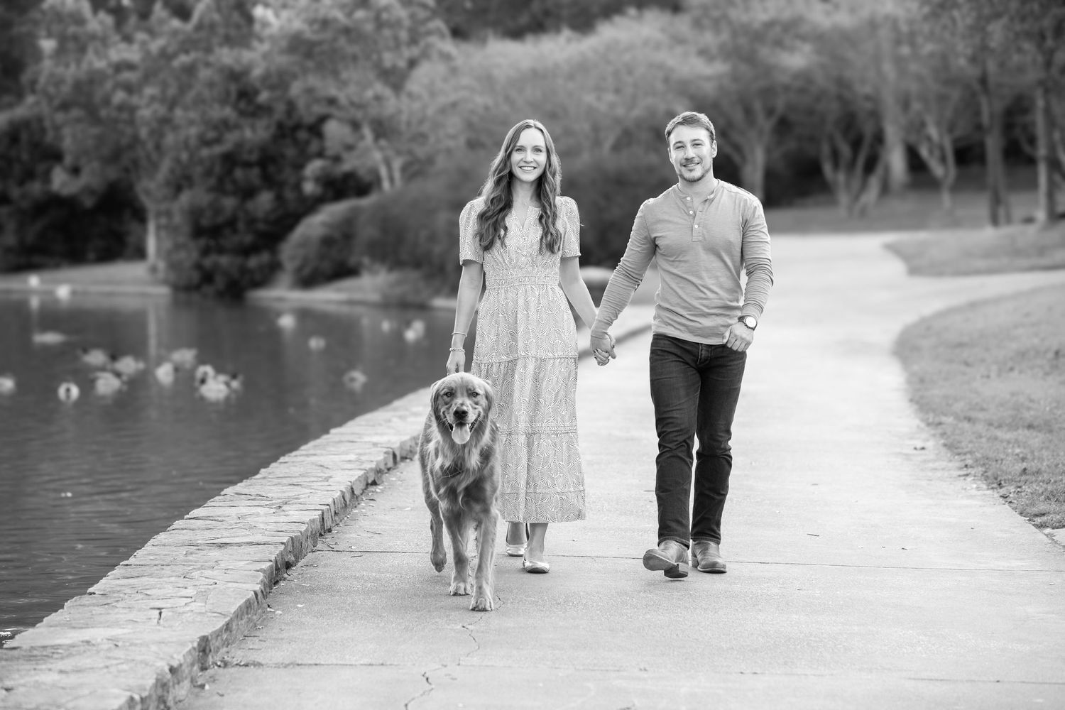 A couple and their dog walk together on a path surrounded by grass and trees in a scenic black and white photo.