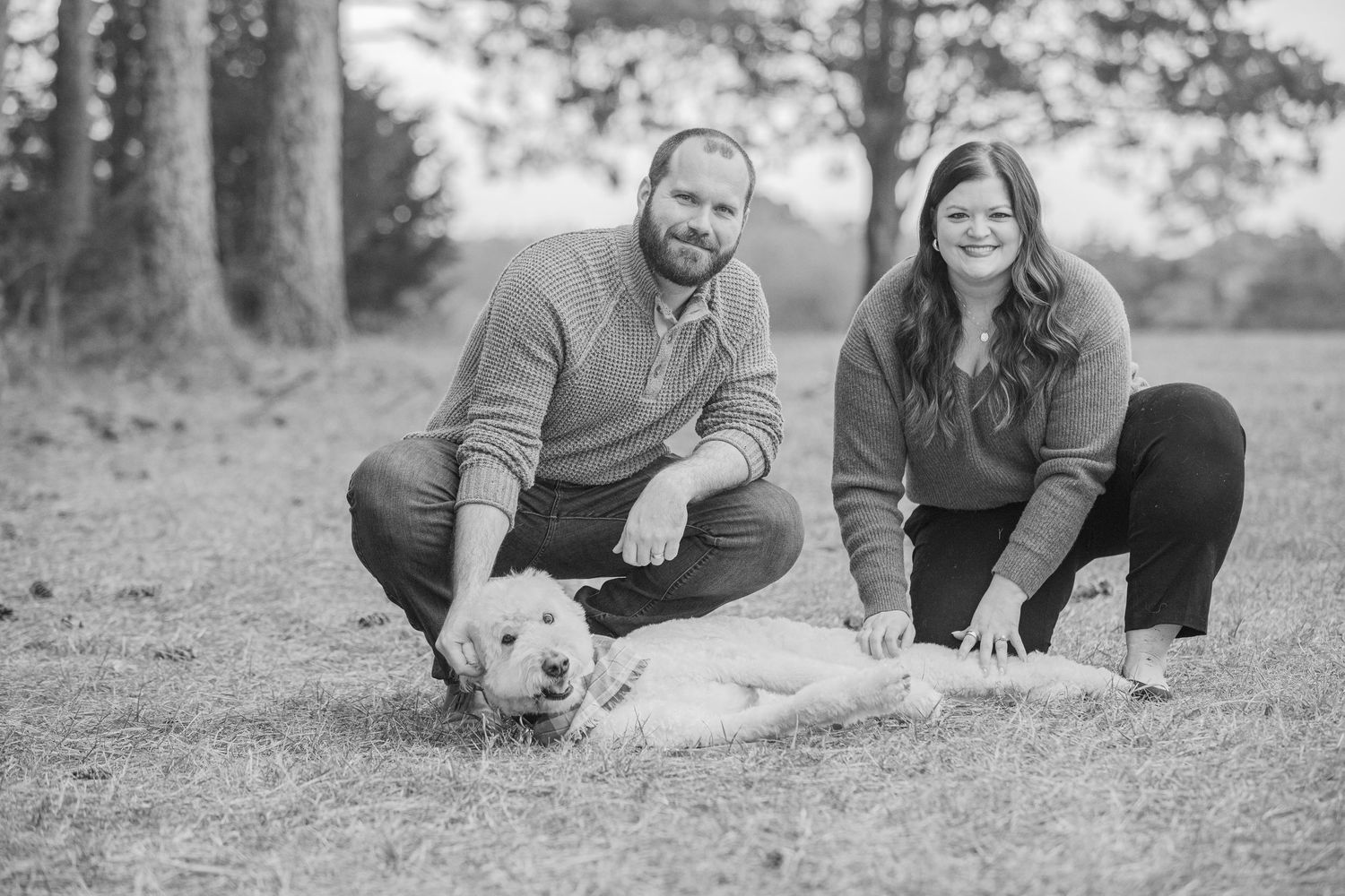Black and white portrait of a couple crouching on grass with their small white dog outdoors.