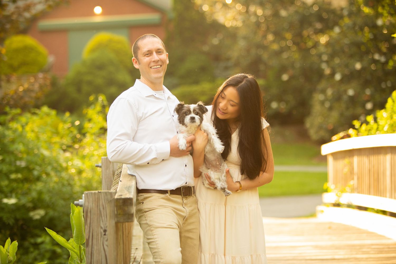 A couple holding their small dog while standing by a wooden fence in a garden at sunset.