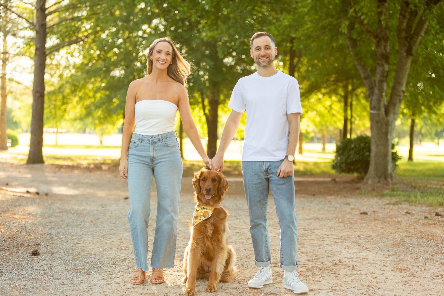 A couple walks with their golden retriever dog along a tree-lined path in warm evening sunlight.