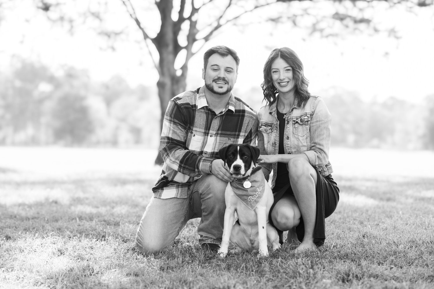 Black and white image of couple kneeling with their dog in an outdoor setting.