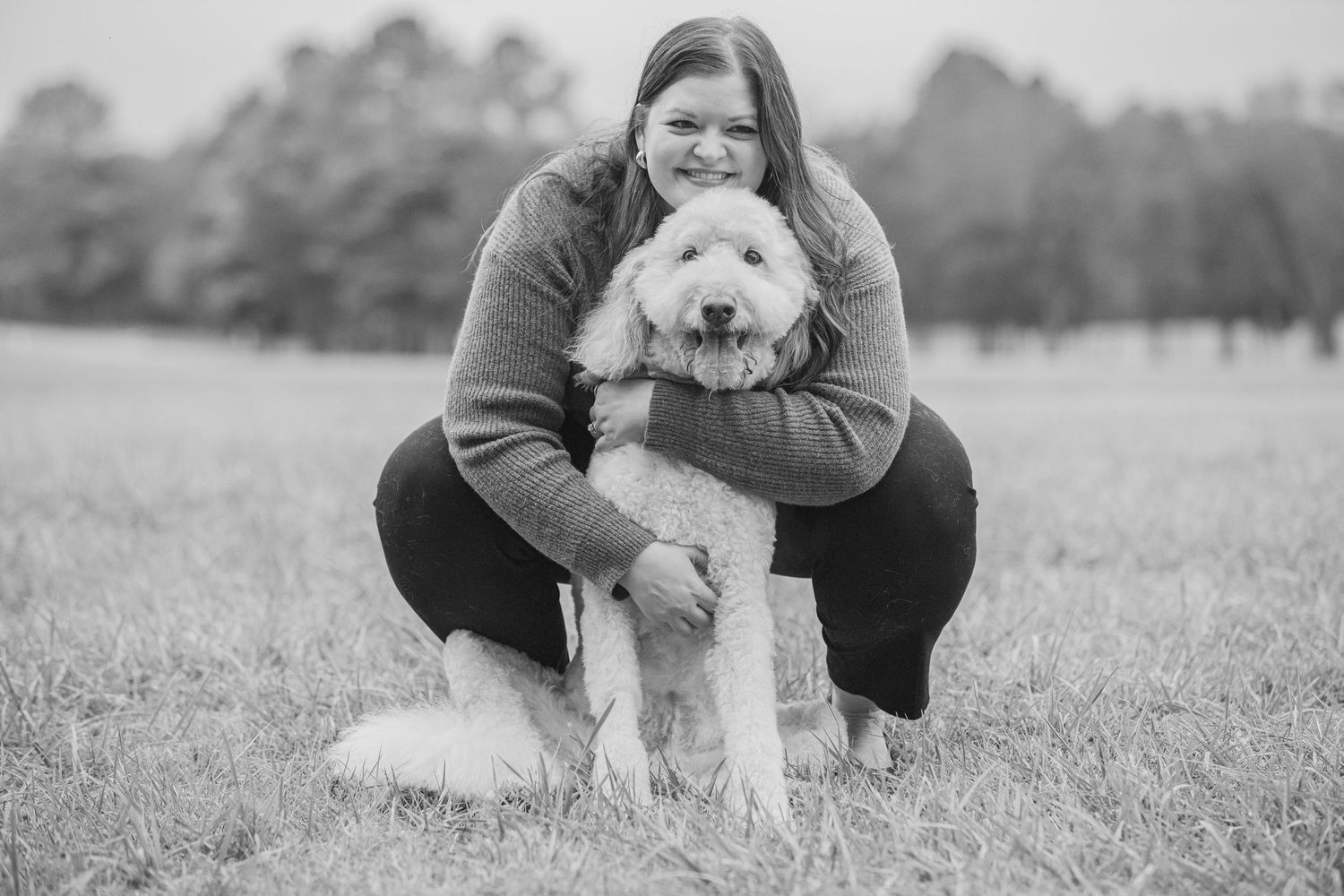Black and white photo of someone crouching in a field while hugging their fluffy white dog.