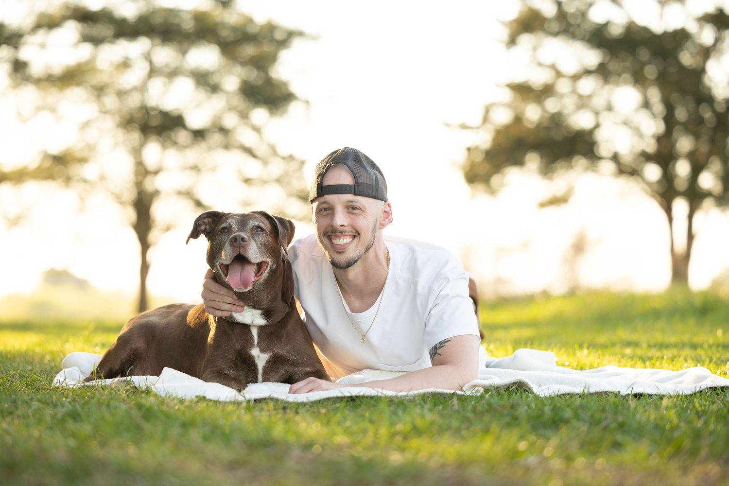 A smiling young man in a white t-shirt and snapback cap lying on a blanket in a park with his brown dog.