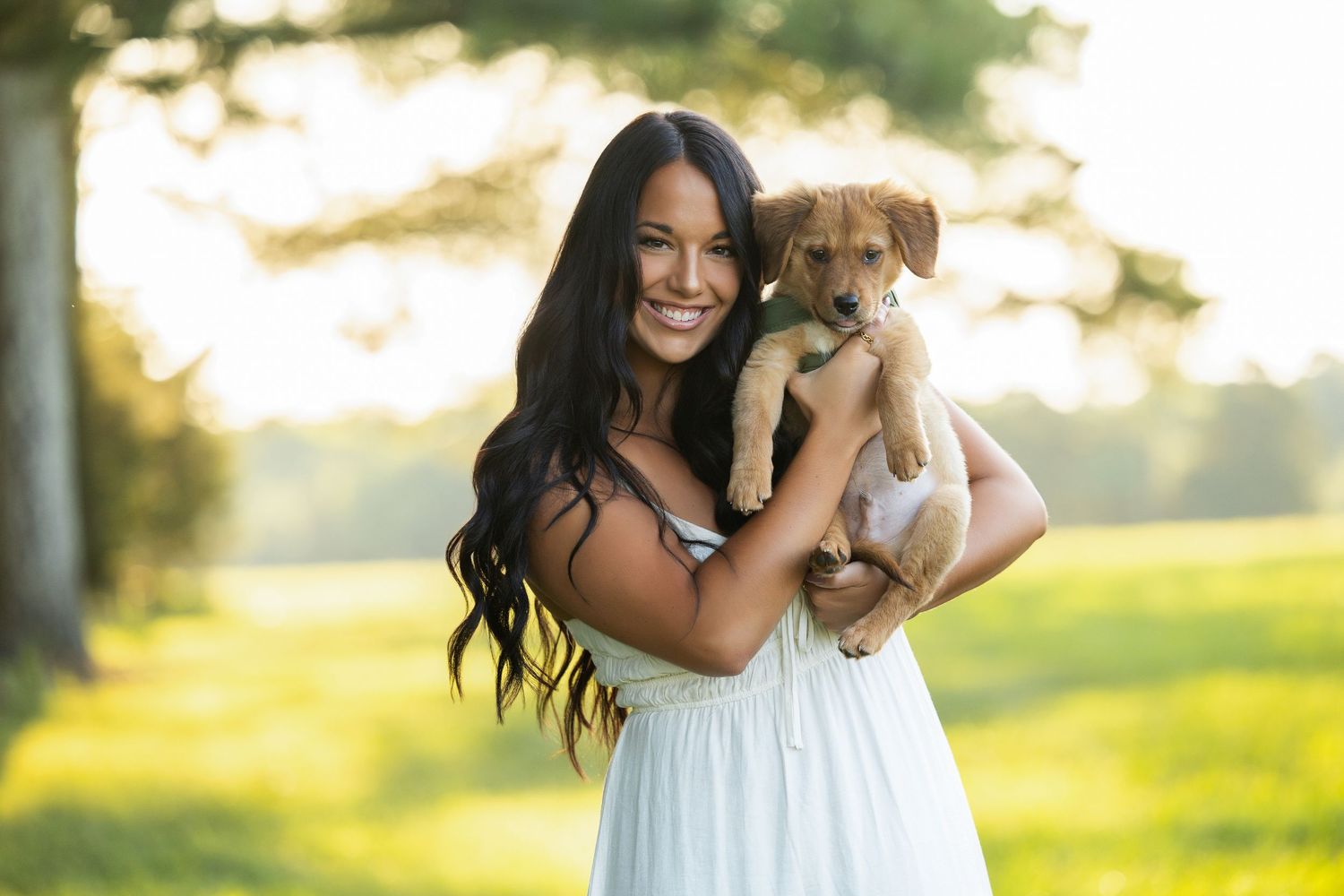 Smiling young woman with long dark hair holding an adorable brown puppy outdoors in a sunny green park.