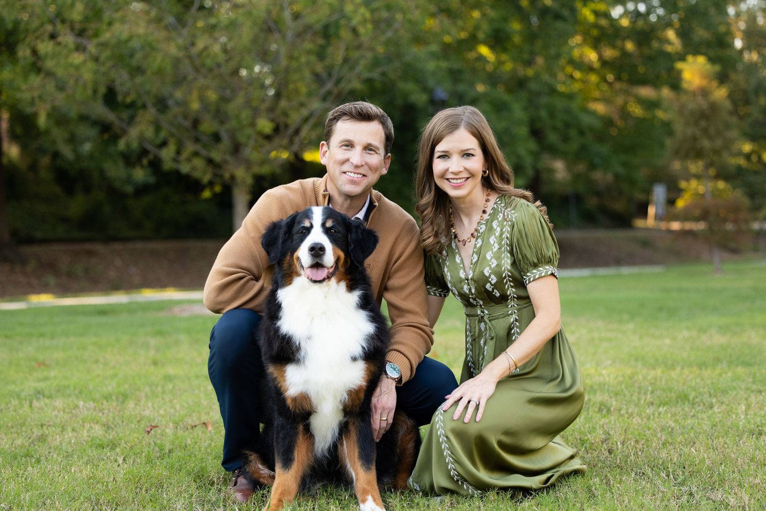 A smiling couple kneels on a grassy lawn in a park posing with their Bernese Mountain Dog on a sunny day.