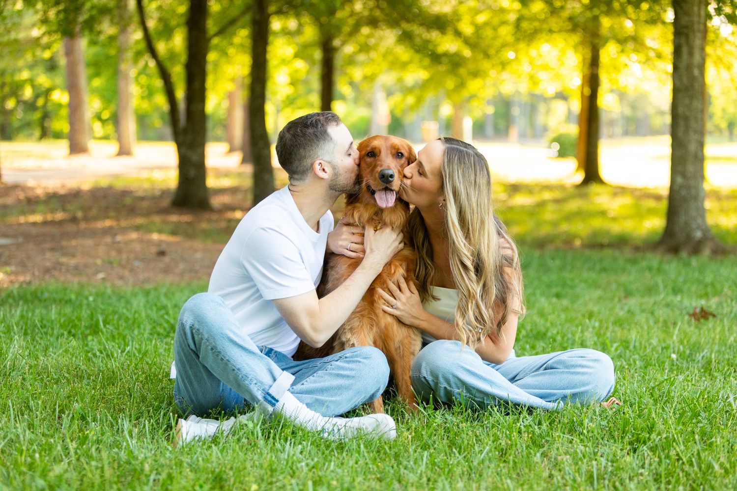 A couple sits on grass in a sunny park while cuddling their golden retriever dog between them.