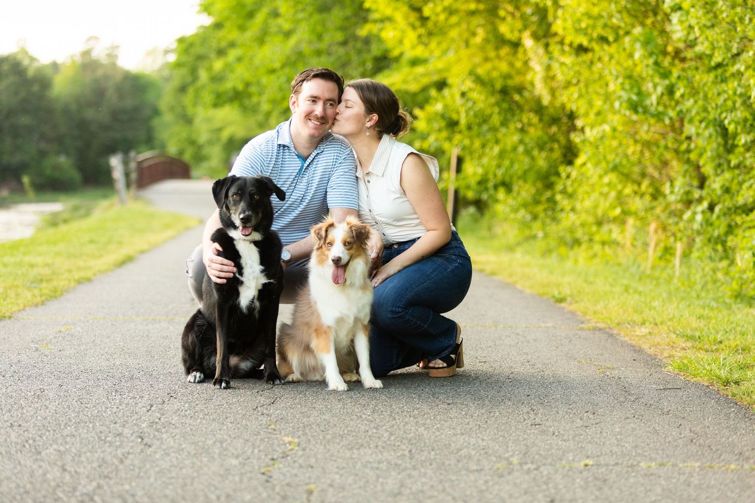 A couple seated on a path outdoors with a black and white dog beside them enjoying a sunny day in nature.