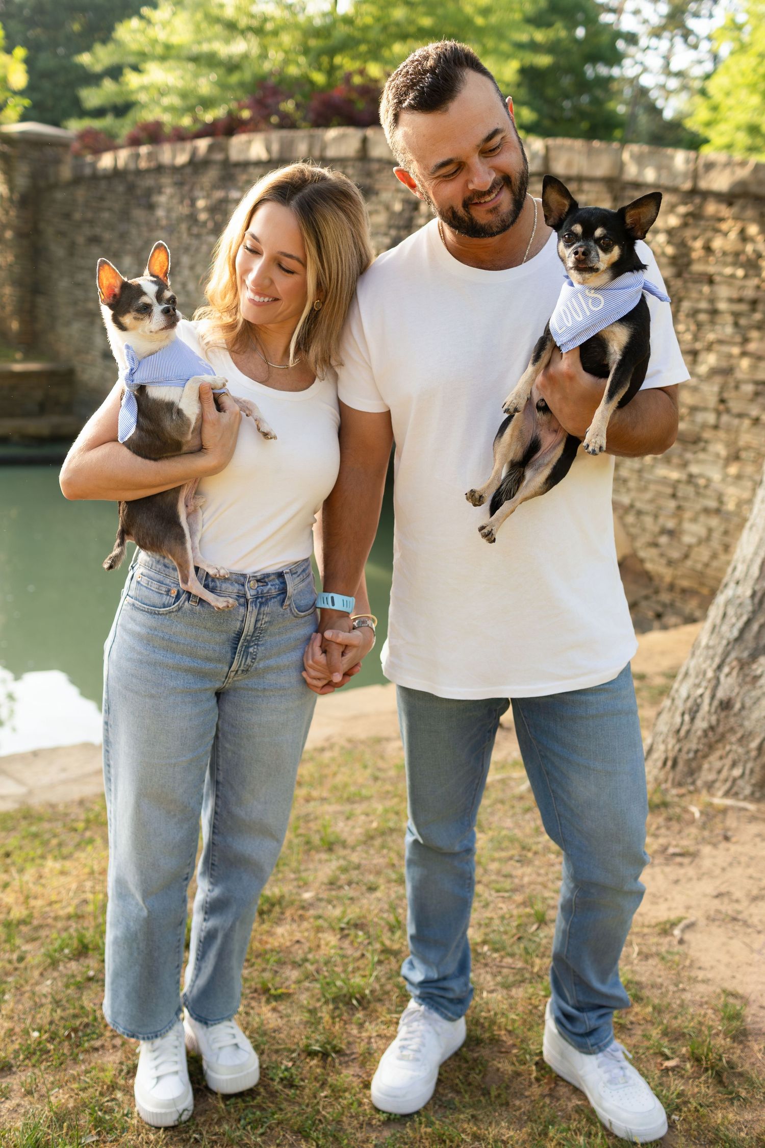 Happy couple in white tees and jeans holding two small Chihuahuas in matching outfits outdoors.
